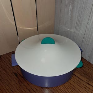 May include: A round purple storage container with a white lid. The lid features a central teal handle, and the container has matching teal side tabs. The design is simple and functional, with a smooth finish on both the base and the lid.