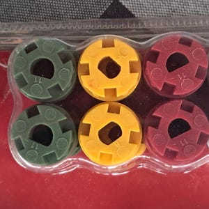 May include: A package of colorful plastic components, including black, green, yellow, red, white, and blue pieces. Each piece has a circular shape with a central hole and a patterned outer edge. The components are arranged in a clear plastic container.
