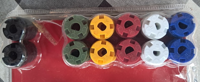 May include: A package of colorful plastic components, including black, green, yellow, red, white, and blue pieces. Each piece has a circular shape with a central hole and a patterned outer edge. The components are arranged in a clear plastic container.