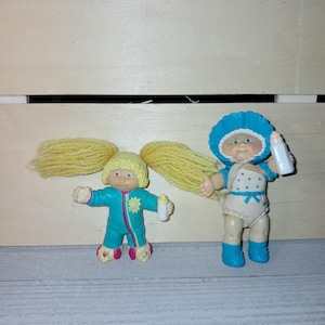 Può includere: Due bambole vintage Cabbage Patch Kids. Una bambola indossa una tuta turchese con dettagli rosa e un fiore giallo. L'altra bambola indossa un cappello blu, un pannolino bianco a pois blu e stivaletti blu. Entrambe le bambole tengono in mano un biberon bianco.