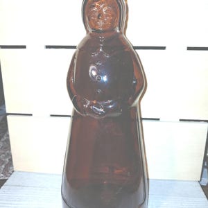 May include: A vintage brown glass bottle shaped like a woman in a long dress. The bottle has a detailed face and a textured surface. The bottle is likely used for storing liquids.