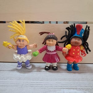 May include: Three Cabbage Patch Kids dolls. The dolls are dressed in different outfits: a ballerina in a purple leotard and white tutu, a girl in a pink dress, and a soldier in a red uniform. Each doll has yarn hair and is holding an accessory.