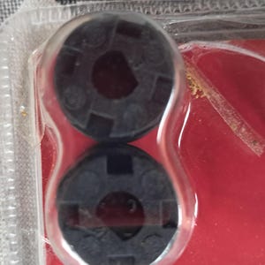 May include: Two black plastic circular objects with a central hole and a patterned surface are encased in clear plastic. The objects are on a red background, suggesting a product package.