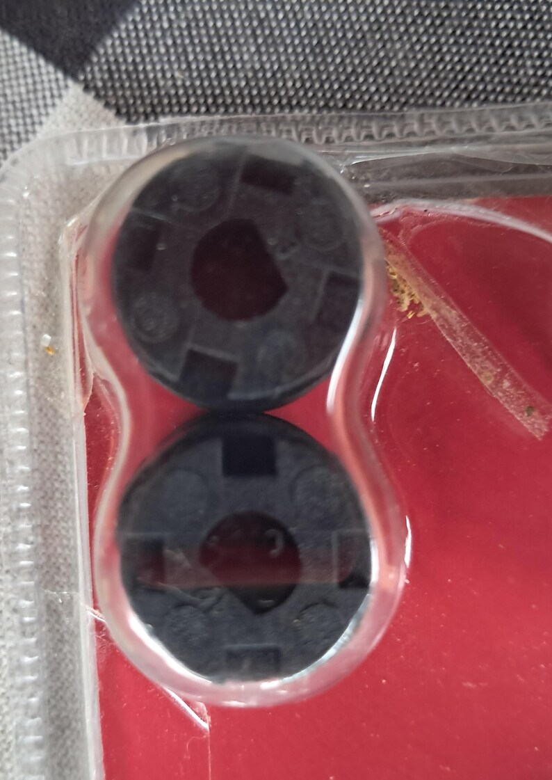 May include: Two black plastic circular objects with a central hole and a patterned surface are encased in clear plastic. The objects are on a red background, suggesting a product package.