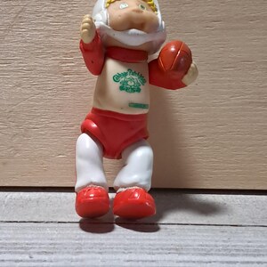 May include: A vintage Cabbage Patch Kids football player doll. The doll is wearing a white helmet, red and white uniform, and holding a small orange football. The doll has a smiling face and the Cabbage Patch Kids logo on the chest.