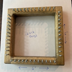 May include: A wooden loom with 12 rows of 12 metal pegs, measuring 6 inches by 6 inches. The loom is made of light brown wood with a darker brown stain. The pegs are silver in color and are evenly spaced around the frame.