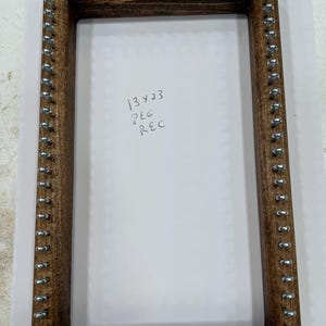 May include: A rectangular wooden weaving loom with metal pegs. The loom is stained a dark brown color and has the text "13 x 23 PEG REC" written on the white surface inside the frame.