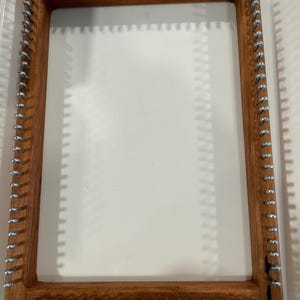 May include: A rectangular wooden weaving loom with metal pegs. The loom is made of light brown wood and is set against a white background. The pegs are evenly spaced around the perimeter of the frame.