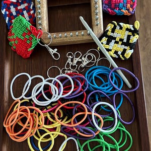 Puede incluir: Un telar de madera con clavijas metálicas, cuadrados tejidos coloridos, llaveros, un ganchillo y bandas elásticas variadas en varios colores. Los artículos están dispuestos en una bandeja de madera, lo que sugiere un tema de artesanía o bricolaje.