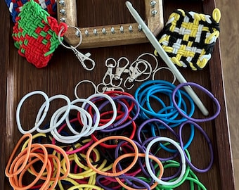 Könnte beinhalten: Ein hölzerner Webrahmen mit Metallstiften, farbige gewebte Quadrate, Schlüsselanhänger, ein Häkelhaken und verschiedene Gummibänder in verschiedenen Farben. Die Artikel sind auf einem Holztablett angeordnet, was auf ein Bastel- oder DIY-Thema hindeutet.