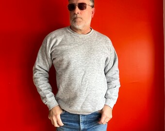 May include: A man wearing a gray sweatshirt and blue jeans. He is standing in front of a solid orange wall and wearing sunglasses.