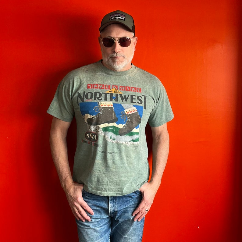 May include: A man wearing a green t-shirt with the text "Take a Hike in the Northwest" and a graphic of hiking boots. He is also wearing a blue baseball cap with the word "Patagonia" on it and blue jeans.