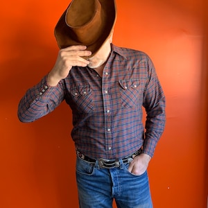 Könnte beinhalten: Ein Mann trägt einen braunen Wildleder-Cowboyhut, ein blau-rotes kariertes Hemd und eine blaue Jeans mit einer silbernen Gürtelschnalle.