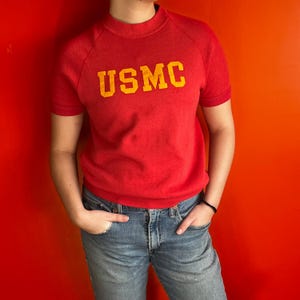 May include: Red short-sleeved sweatshirt with the yellow text "USMC" across the chest. The shirt has a crew neck and raglan sleeves. The person is wearing blue jeans.