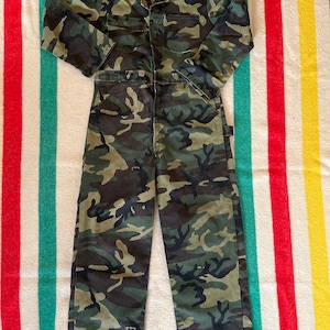 Könnte beinhalten: Ein Overall mit langem Arm in Tarnmuster mit Reißverschluss. Der Overall hat eine große Tasche auf der Brust und zwei kleinere Taschen an den Hüften.