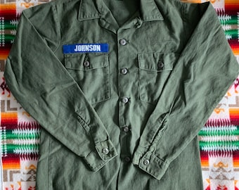 May include: A green long-sleeved shirt with a blue patch that says "JOHNSON" on the left chest. The shirt has two flap pockets and a button-up front.