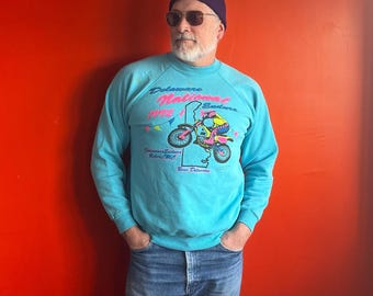 May include: A turquoise blue sweatshirt with a graphic print of a motocross rider jumping over a ramp. The graphic includes the text "Delaware National Enduro 1992" and "Delaware Enduro Riders MC". The person wearing the sweatshirt is wearing a black beanie and sunglasses.