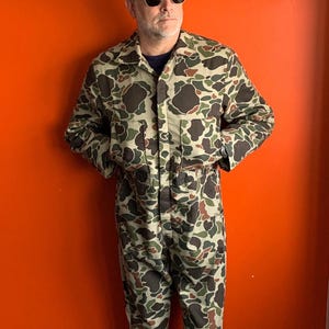 Puede incluir: Una persona con un mono de camuflaje, una boina naranja brillante y gafas de sol. La persona está de pie frente a un fondo naranja sólido.