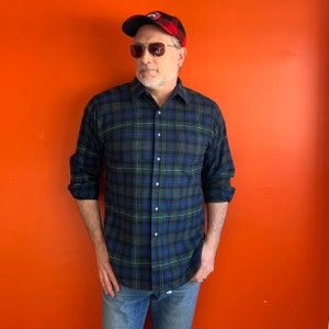 May include: A man wearing a blue and green plaid long-sleeved shirt with a red and black plaid baseball cap. He is wearing blue jeans and sunglasses.