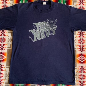May include: A navy blue t-shirt with a white graphic of the words "THE NAVY" in a 3D block style with an eagle and anchor emblem above the words.