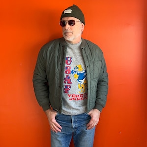 May include: A man wearing a green quilted jacket, a grey sweatshirt with a cartoon eagle graphic and the text "USAF Yokota Japan", and a dark green beanie with a brown brim. He is wearing blue jeans and brown sunglasses.