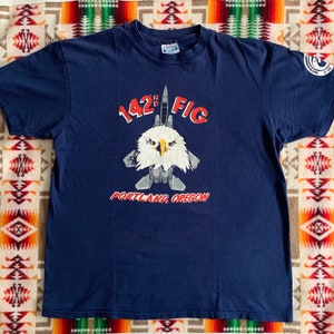 May include: A navy blue t-shirt with a graphic of a fighter jet and an eagle head. The text "142nd FIG PORTLAND, OREGON" is printed on the shirt.