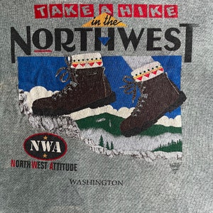 May include: A gray t-shirt with a graphic design of hiking boots and a mountain range. The text "TAKE A HIKE in the NORTHWEST" is printed in black letters. The text "NWA NORTHWEST ATTITUDE" is printed in black letters with red stars. The text "WASHINGTON" is printed in black letters.