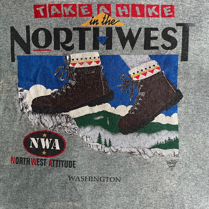 May include: A gray t-shirt with a graphic design of hiking boots and a mountain range. The text "TAKE A HIKE in the NORTHWEST" is printed in black letters. The text "NWA NORTHWEST ATTITUDE" is printed in black letters with red stars. The text "WASHINGTON" is printed in black letters.