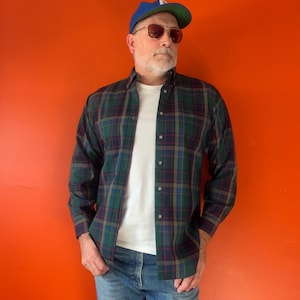 May include: A long-sleeve button-down shirt featuring a dark green, navy blue, and maroon plaid pattern. The shirt is worn over a plain white t-shirt, paired with blue denim jeans, a blue baseball cap, and tinted sunglasses with thin metal frames.