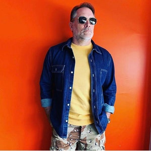 May include: A man wearing a blue denim shirt over a yellow t-shirt and camouflage cargo pants. He is wearing sunglasses and has a beard.