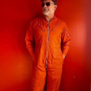 May include: A man wearing a bright orange jumpsuit, a brown baseball cap, and sunglasses stands in front of a red wall.