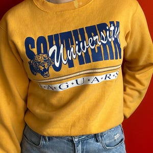 Peut inclure: Sweat-shirt jaune moutarde avec l'inscription "Southern University" en bleu marine et blanc. Le mot "Jaguars" est en dessous en blanc. Un motif de tête de jaguar est sur le côté gauche du sweat-shirt.