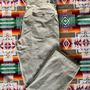 May include: A pair of beige utility pants with a large cargo pocket on the back. The pants are folded and laid flat on a patterned blanket.