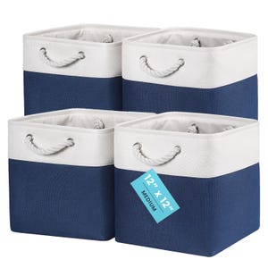 May include: A set of four square storage bins featuring a two-tone design with a dark blue base and a white top section. Each bin includes white rope handles attached with silver metal grommets. One bin displays a blue tag reading 12" x 12" MEDIUM.