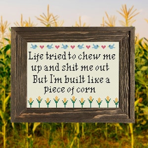 Op de afbeelding: Een ingelijst kruissteekontwerp met een witte achtergrond en een bruin frame. Het ontwerp bevat een citaat: "Life tried to chew me up and shit me out But I'm built like a piece of corn." Het citaat is omringd door blauwe vogels en roze harten. De onderkant van het ontwerp bevat een rij gele maïs.