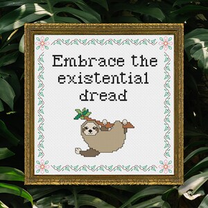 May include: Cross-stitch art in a gold frame. The artwork features the text "Embrace the existential dread" above a cartoon sloth hanging from a branch. The frame has a floral and vine border.