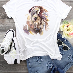 May include: White t-shirt with a watercolor painting of a brown horse's head. The horse has a long mane and is looking down. The t-shirt is paired with denim shorts with star-shaped embellishments.