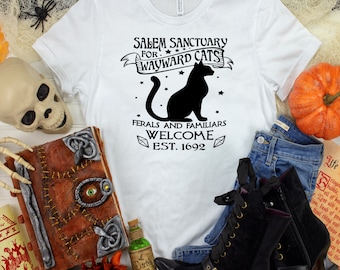 May include: White t-shirt with black text that reads "Salem Sanctuary for Wayward Cats" with a black silhouette of a cat and stars. The text below the cat reads "Feral and Familiars Welcome Est. 1692".
