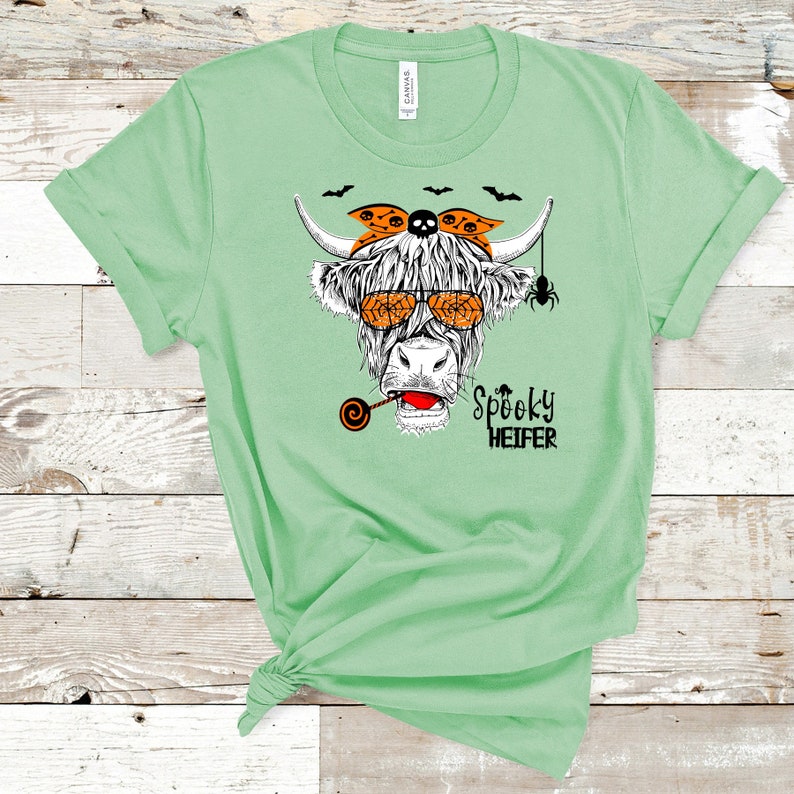K&ouml;nnte beinhalten: Ein gr&uuml;nes T-Shirt mit einer Schwarzwei&szlig;-Illustration einer Kuh, die eine Sonnenbrille und ein Bandana mit Totenkopf und Knochen tr&auml;gt. Die Kuh h&auml;lt einen Lolli im Maul. Der Text "Spooky Heifer" ist unter der Kuh aufgedruckt.