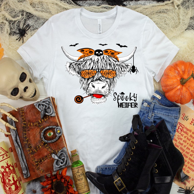 K&ouml;nnte beinhalten: Wei&szlig;es T-Shirt mit einem Grafikdruck einer schottischen Hochlandkuh, die eine Sonnenbrille, ein Bandana und einen Lolli tr&auml;gt. Die Kuh hat einen gruseligen Ausdruck und der Text "Spooky Heifer" ist unter dem Bild gedruckt.