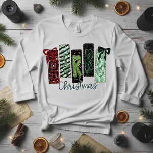 Puede incluir: Camiseta blanca de manga larga con un gráfico de "Merry Christmas". El diseño presenta formas rectangulares coloridas que se asemejan a regalos, adornadas con lazos y la palabra "Merry" en verde. La palabra "Christmas" está en cursiva.
