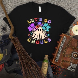 Puede incluir: Camiseta negra con un fantasma blanco que lleva una corona de flores y signos de paz. El texto "Let's Go Ghouls" est&aacute; en colores arco&iacute;ris.