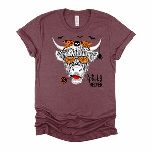K&ouml;nnte beinhalten: Ein maroonfarbenes T-Shirt mit einer wei&szlig;en Hochlandkuh, die eine orangefarbene Sonnenbrille und ein Bandana mit Totenkopf und Knochen tr&auml;gt. Die Kuh h&auml;lt einen Lolli und &uuml;ber ihrem Kopf befindet sich eine Spinne. Der Text "Spooky Heifer" ist unter der Kuh aufgedruckt.
