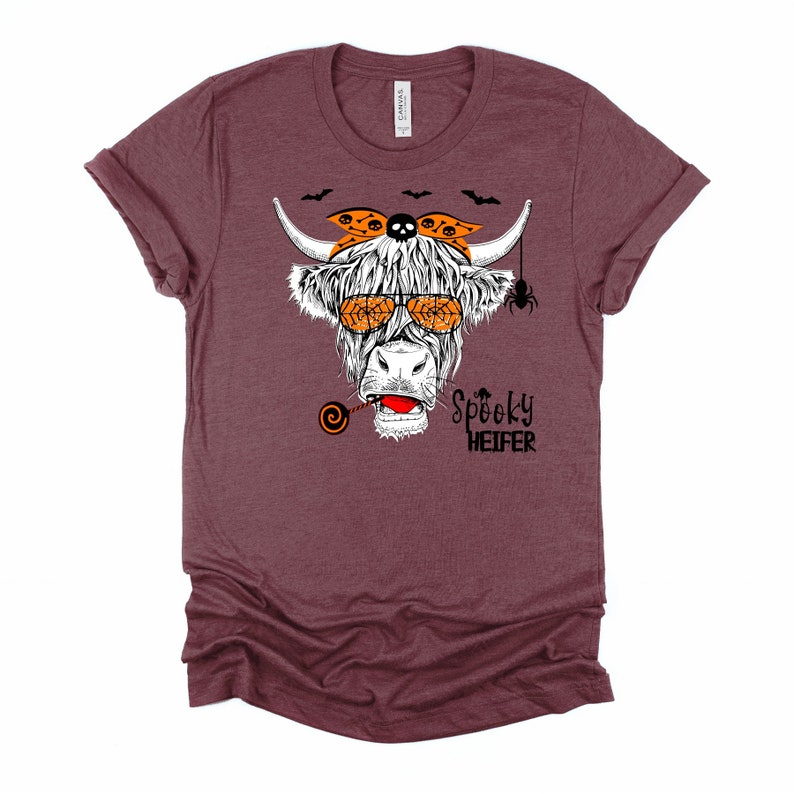 K&ouml;nnte beinhalten: Ein maroonfarbenes T-Shirt mit einer wei&szlig;en Hochlandkuh, die eine orangefarbene Sonnenbrille und ein Bandana mit Totenkopf und Knochen tr&auml;gt. Die Kuh h&auml;lt einen Lolli und &uuml;ber ihrem Kopf befindet sich eine Spinne. Der Text "Spooky Heifer" ist unter der Kuh aufgedruckt.
