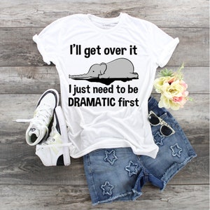 Könnte beinhalten: Weißes T-Shirt mit schwarzem Text, der "I'll get over it I just need to be DRAMATIC first" lautet. Ein grauer Elefant aus Zeichentrick mit einem langen Rüssel liegt auf dem T-Shirt.