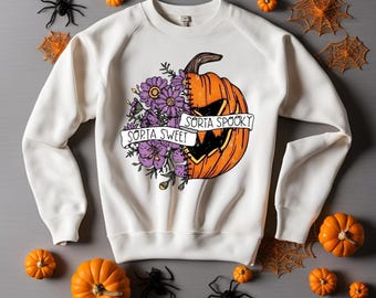 May include: White sweatshirt with a Halloween-themed graphic. The design features a pumpkin with a floral arrangement and the text "Sorta Spooky" and "Sorta Sweet". The image is decorated with pumpkins, spiders, and spiderwebs.