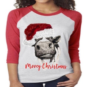 Peut inclure: Un t-shirt de style baseball blanc et rouge avec une vache portant un chapeau de Père Noël et le texte "Mooey Christmas".