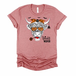 K&ouml;nnte beinhalten: Ein pinkfarbenes T-Shirt mit einer Schwarzwei&szlig;illustration einer Kuh, die eine Sonnenbrille, ein Bandana und einen Lutscher tr&auml;gt. Der Text "Spooky Heifer" ist unter der Kuh aufgedruckt.