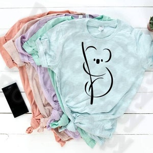 Könnte beinhalten: Ein Stapel aus fünf T-Shirts in verschiedenen Pastellfarben, darunter hellrosa, Lavendel, Mintgrün und hellblau. Das oberste Shirt ist hellblau und zeigt eine schwarze Linienzeichnung eines Koalabären, der sich an einem Ast festhält.