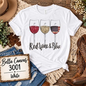Könnte beinhalten: Ein weißes Kurzarm-T-Shirt mit einer Grafik von drei Weingläsern, gefüllt mit rotem Glitzer, goldenem Glitzer und einem US-Flaggenmuster, mit dem Text Red Wine & Blue in schwarzer Schreibschrift unter den Gläsern.
